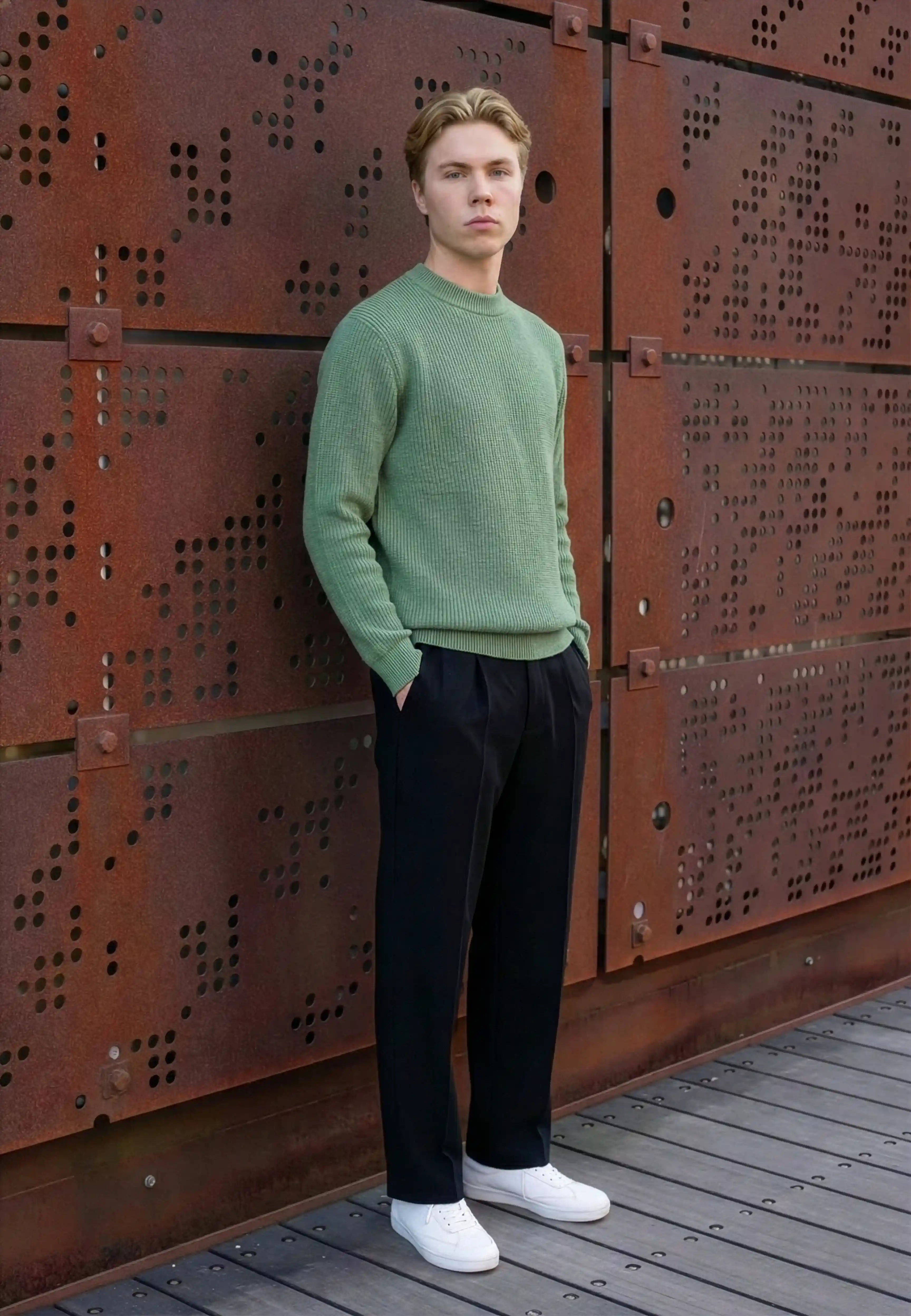 Man in a green knitted sweater and black trousers against a rust coloured steel wall