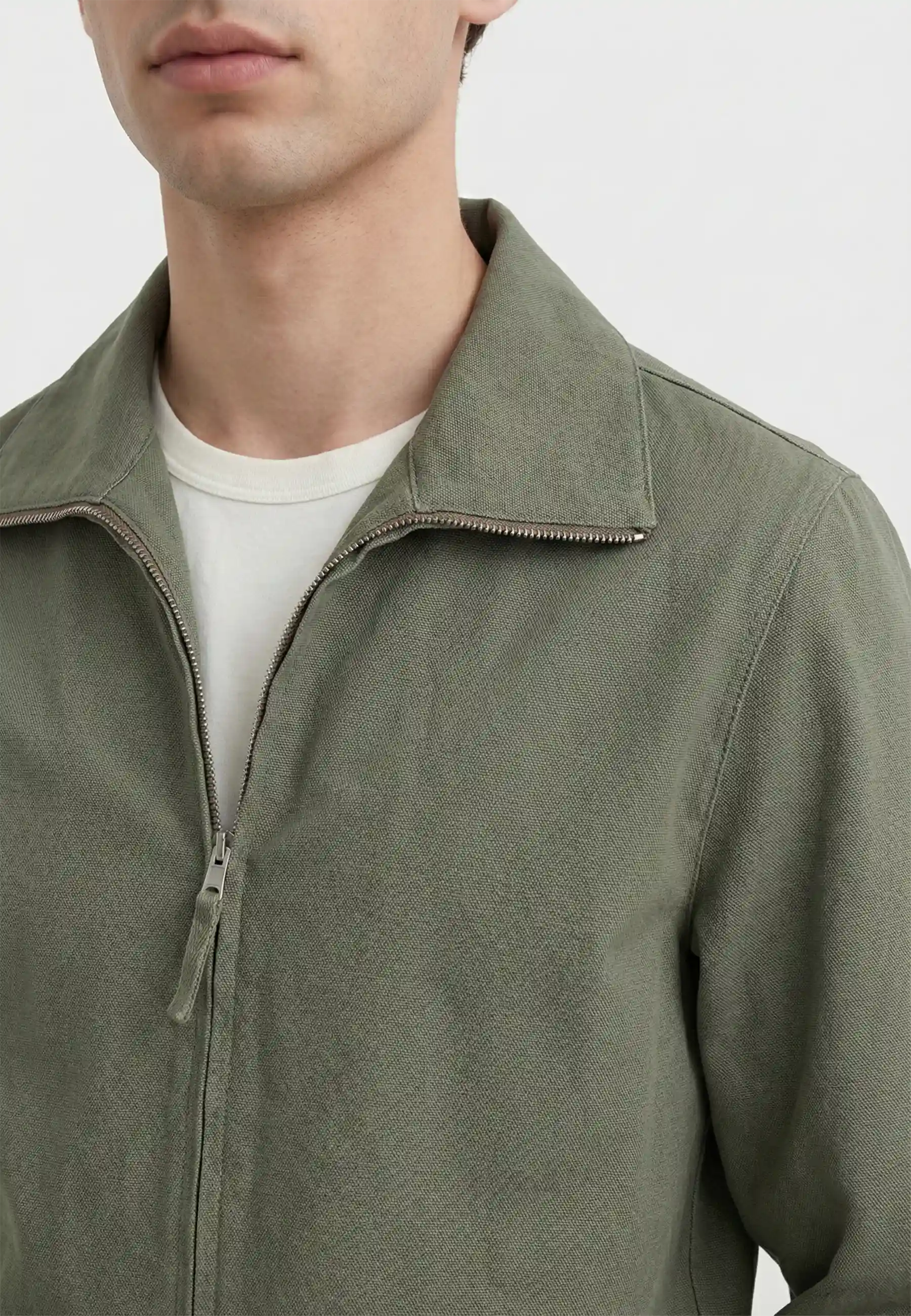 Närbild på en olivgrön zip-up jacka i textil med krage och dragkedja, buren över en vit t-shirt på en man.