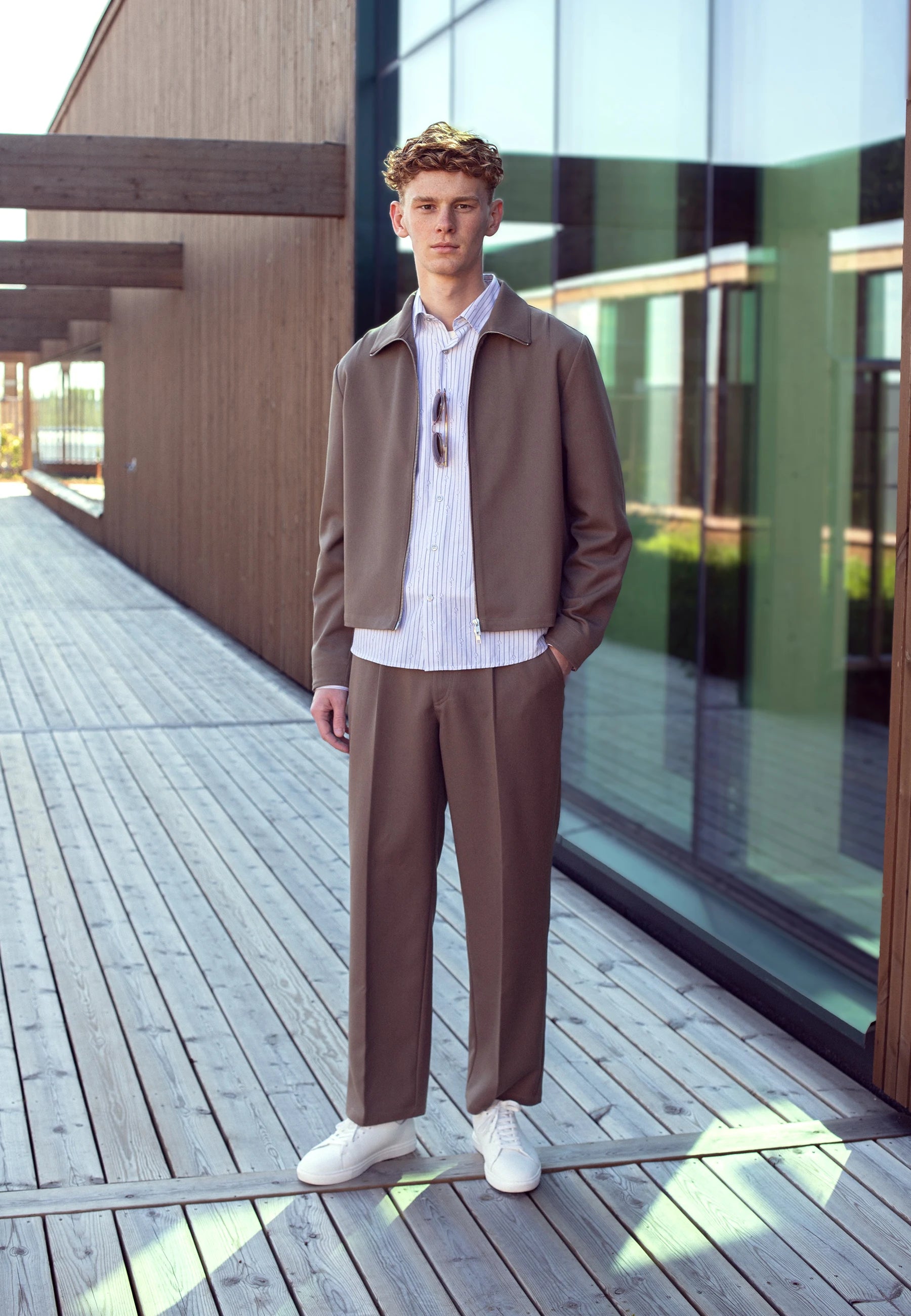 Man in a brown jacket and pants standing on a wooden deck with modern architecture in the background